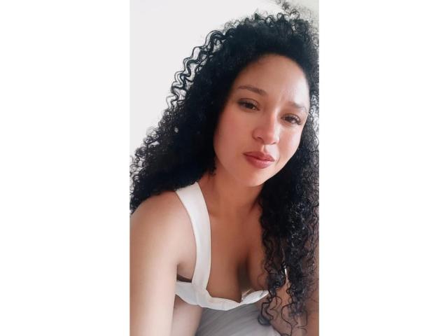 CarlaMuille with curly black hair, light brown skin, wearing a white tank top, showing her small tits, and giving a slight pout, looking directly at the camera.