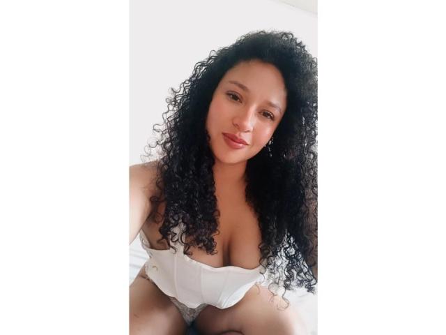 CarlaMuille with curly black hair, light brown skin, and red lipstick, wearing a white, low-cut top, showing her tits, sitting on a bed, looking at the camera.
