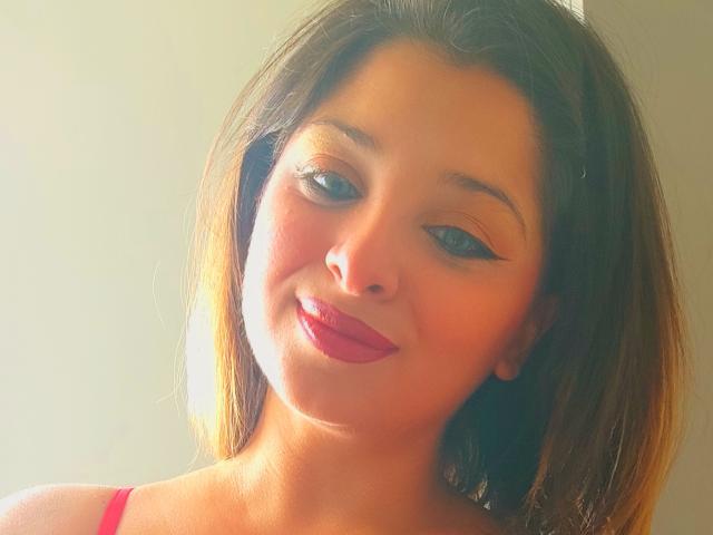 ScarletOra hat eine mittelbraune Haut, dunkelbraunes, schulterlanges Haar und roten Lippenstift mit schwarzem Eyeliner. Sie lächelt und das Sonnenlicht scheint auf ihr Gesicht. Ihre Brüste sind klein und niedlich..