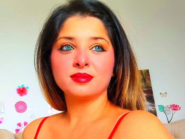 ScarletOra heeft blauwe ogen, rode lippenstift en zwart haar met blonde highlights, dat halverwege haar schouders komt. Ze draagt een rode top en kijkt recht in de camera met een lichte glimlach..