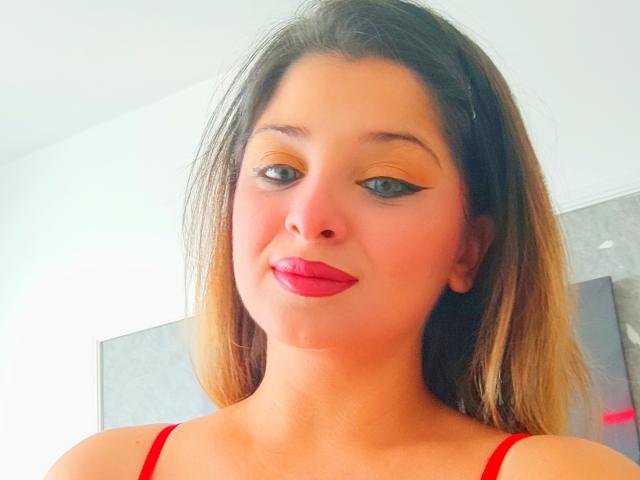 ScarletOra met een lichtbruine huid, zwarte eyeliner, oranje oogschaduw, rode lipstick en steil blond haar, draagt een rode top en glimlacht lichtjes..