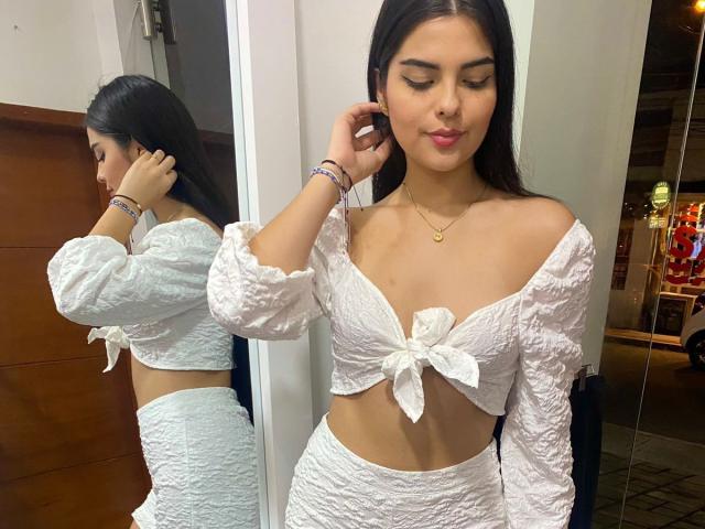 NicoleHarris with long black hair, wearing a white, textured, off-shoulder crop top tied in front, and matching high-waisted skirt, holding her ear, reflected in a mirror.