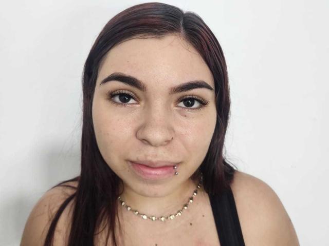 A AniHotty tem olhos castanho-escuros, cabelo preto-avermelhado liso, um piercing no nariz, um piercing no lábio e veste uma regata preta com um colar de contas douradas..
