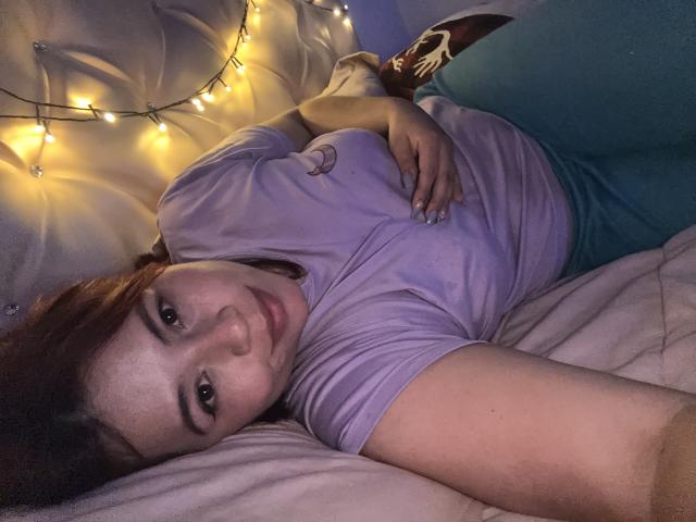 MelisaBrunette is lying on a bed, wearing a purple shirt and blue pants, with a relaxed expression, hands on her chest, and a string of yellow lights in the background.