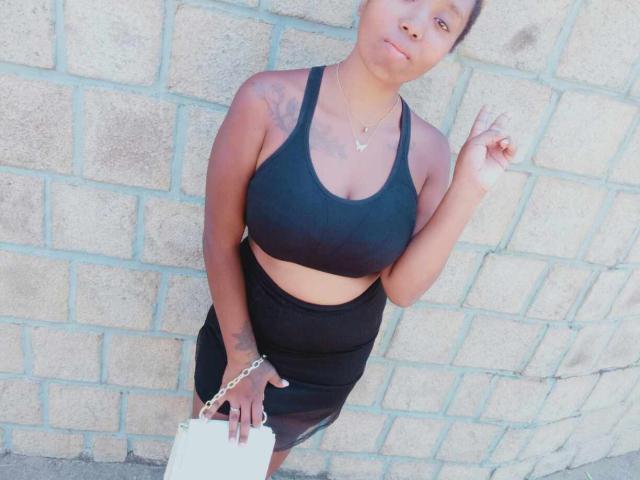 Jobba has medium brown skin, black sports bra showing her tits, black high-waisted skirt, white handbag, holding finger up, short curly hair, necklace, standing against brick wall.
