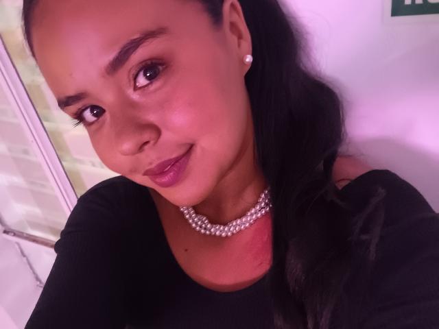 ArielleWilde with dark skin, black hair in a ponytail, wearing a pearl necklace and black top, smiling with pinkish-purple lipstick. Boobs not visible. Her face is centered.