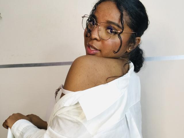 Lisa Mendez, de piel oscura, cabello negro rizado y gafas, vestida con una camisa blanca con un hombro descubierto, mirando por encima del hombro con lápiz labial rosa, mostrando su hombro descubierto y un tatuaje..