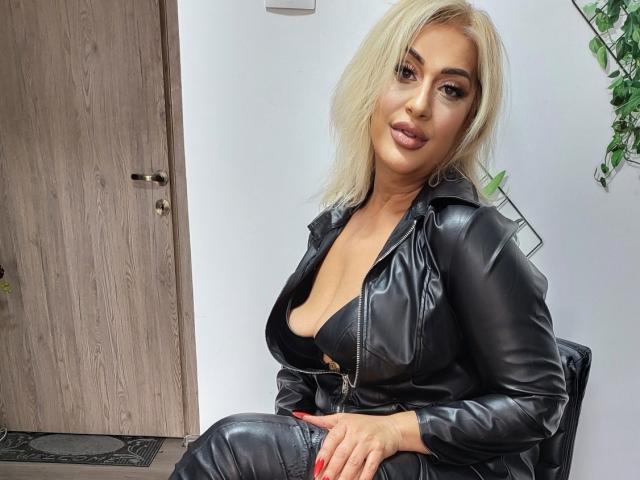 Una donna attraente con capelli biondi, seno prosperoso, indossa una giacca di pelle nera con zip, è seduta, guarda la fotocamera, ha le unghie rosse, sullo sfondo si vede una porta di legno, a destra c'è una pianta, appare sicura di sé e sexy..