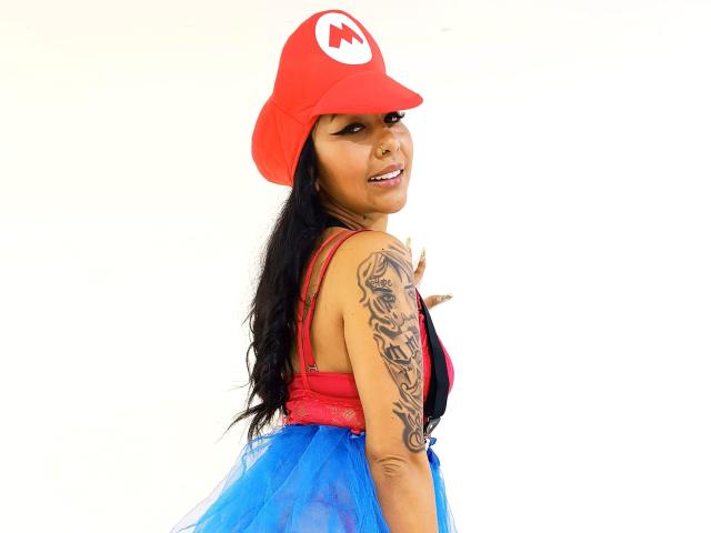 TracyLambo with a red cap, long black hair, tattooed arm, red bra, and blue tulle skirt, looking back with a smile. Boobs visible, cap has a white "M" logo.