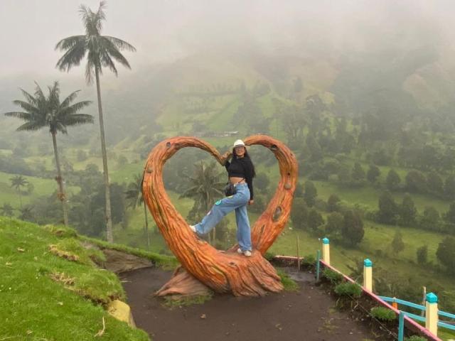 ChloeLauras standing on a wooden heart sculpture, wearing a black top, blue jeans, and white beanie, with foggy hills and palm trees in the background.