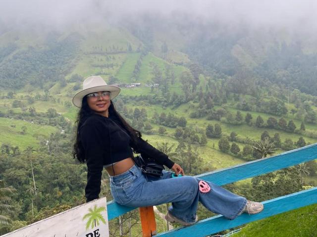 ChloeLauras with long black hair, wearing a white hat, black crop top, blue jeans, and pink socks, sitting on a blue railing, showing her ass, in a foggy mountain.