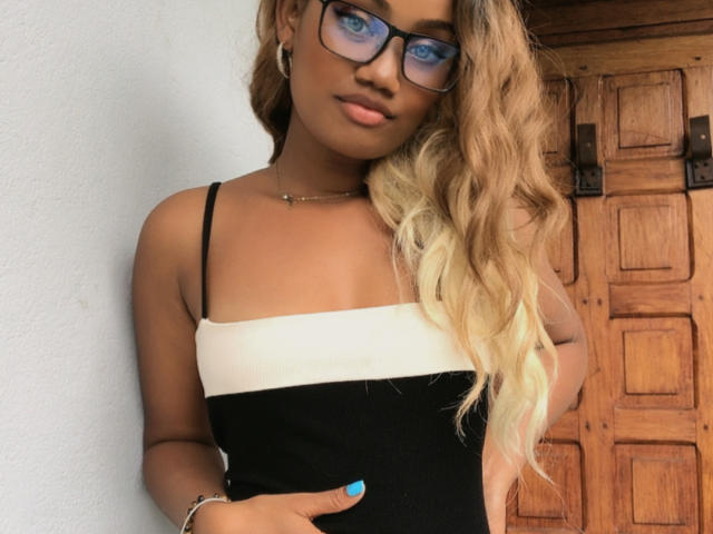 Loyrianah with wavy blonde hair, black and white dress, blue nails, glasses, and bracelet, standing against a white wall, hand on hip, wooden door in background.