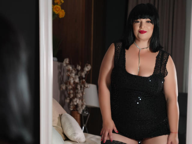 BustyRubi stands in a mirror, wearing a black lace dress, showing her big boobs, with a silver necklace, black bob haircut, and red lipstick, in a softly lit room.