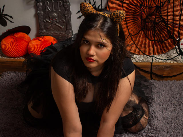 ShadowBlue, con cabello oscuro, orejas de leopardo, vestido negro y medias de red, a cuatro patas, mirando a la cámara, con lápiz labial rojo y cicatrices en la mejilla, sobre una alfombra gris..