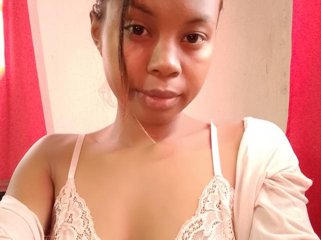 Marielie with brown skin, small boobs in a white lace bra, and a subtle smile, wearing a pink bra strap and a white shirt, in a selfie.