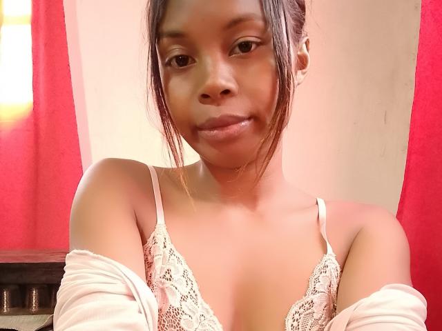 Marielie with medium brown skin, dark hair in a ponytail, wearing a lace bra, white off-shoulder top, and small earrings, smiling slightly at the camera.
