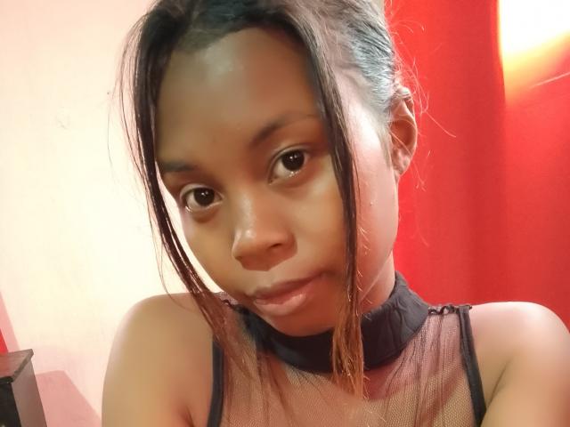 Marielie has dark skin, brown eyes, and medium brown hair in a messy updo. She wears a black mesh top, showing her small, bare shoulders and a subtle smile.