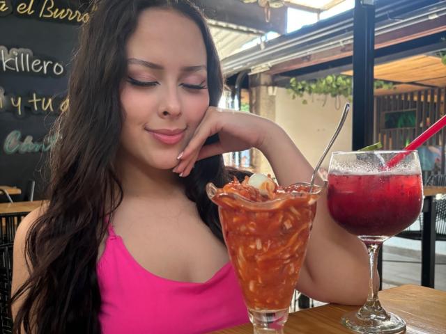AlisonTatcher with long dark hair, pink tank top, and pink lips, smiling, resting chin on hand, holding a tall glass of spaghetti ice cream, and a red drink.