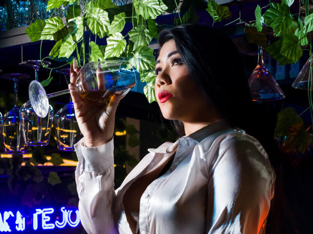 KhloeLawson holding a glass of amber liquid, wearing a white button-up shirt, with dark hair and red lipstick, looking up with a slight smile. Boobs slightly visible under shirt.