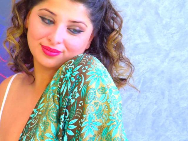 ScarletOra, con cabello castaño rizado, un chal de color verde y dorado con estampado floral, lápiz labial rosa y sombra de ojos azul, sonriendo a la cámara, mostrando su top con tirantes blancos y escote..