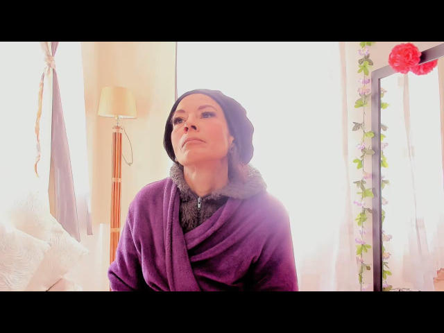 TatianaPotter69 wearing a purple shawl and black beanie, looking up with a slightly annoyed expression, soft sunlight on her face, standing in a cozy room.
