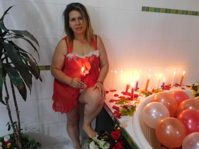 SevageLove, middle-aged with brown hair, red lace lingerie, standing in a bathroom, one leg up, holding a candle, next to a candlelit, rose-adorned bathtub with balloons.