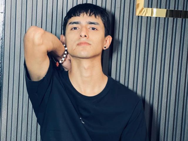 NathanMaxime, avec ses cheveux noirs courts, son t-shirt noir et ses boucles d'oreilles noires, bras levé, l'air légèrement ennuyé, portant un bracelet de perles blanc et rouge..
