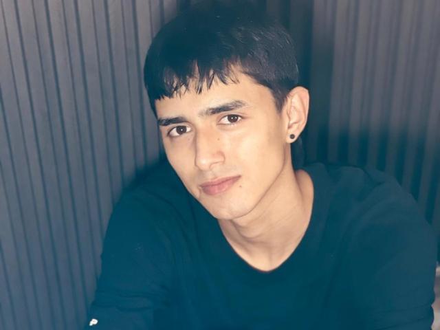 NathanMaxime, a cute young dude with short black hair, black shirt, and black ear stud, smiling softly at the camera, showing a smooth, slightly freckled face.