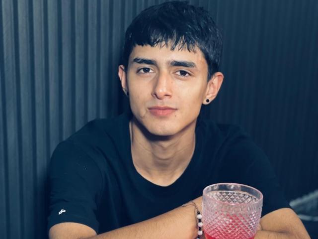 NathanMaxime, con cabello negro corto, camiseta negra, un pequeño agujero en la oreja, sosteniendo un vaso de vidrio con un estampado de diamantes rosas, con una expresión ligeramente aburrida, de pie frente a una pared oscura con textura..