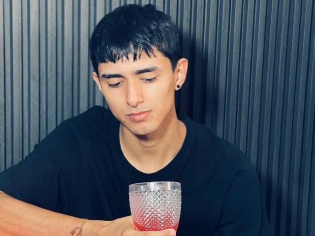 NathanMaxime, met kort zwart haar, een zwart T-shirt, die een geribbeld rood glas vasthoudt, naar beneden kijkend, met een kleine oorbel, op een donkere achtergrond met verticale lijnen..