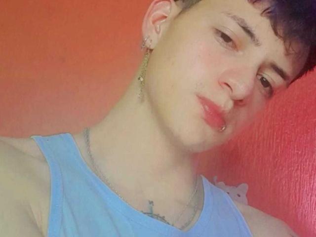 GabyThoms has a short, dark haircut, wearing a light blue tank top, nose piercing, lip ring, and a cross tattoo. She's looking to the side with a slight pout.