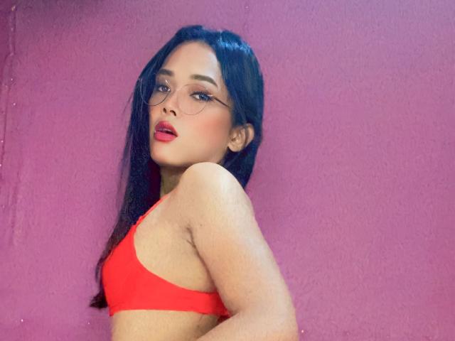 SpicyInfinity, avec ses longs cheveux noirs, son rouge à lèvres rouge et ses lunettes, porte un débardeur de sport rouge, montrant ses seins contre un mur rose. Elle regarde par-dessus son épaule avec un regard sexy..
