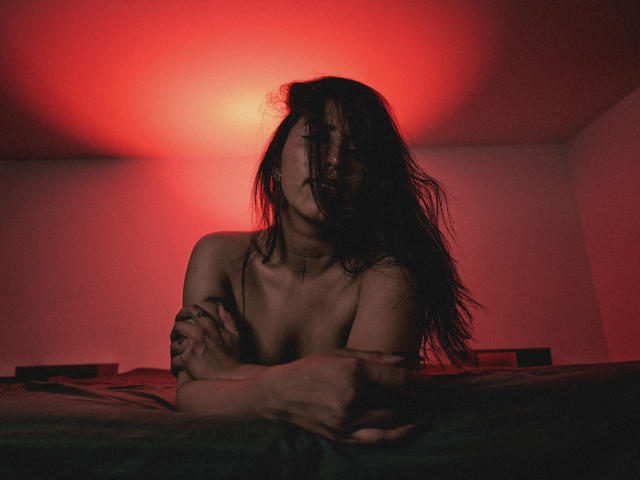 ElizabethMacy, naked, with long dark hair, arms crossed, small tits, red light, looking down, dark shadows, bed in foreground, serious expression, moody atmosphere.