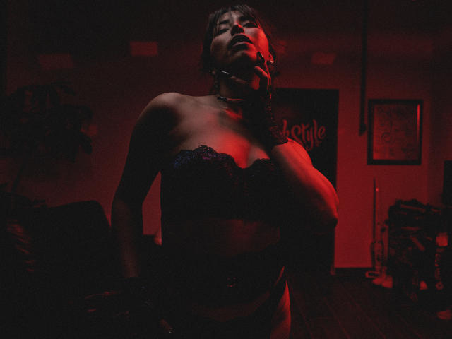 Elizabeth Macy met een zwarte kanten BH, rode verlichting, een choker, en los haar, haar borsten prominent, hand in de buurt van haar nek, naar boven kijkend, in een zwak verlichte, sexy kamer..