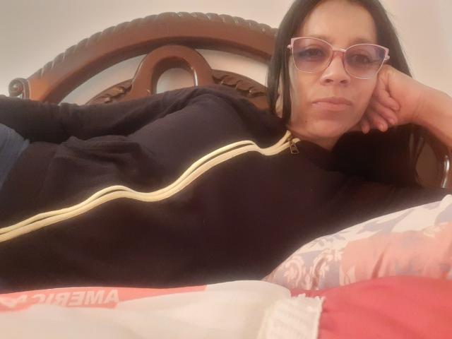 IsabellaSweett is lying on a bed, wearing a black zip-up jacket with gold stripes, and pink glasses, with her head resting on her hand, showing her ass.