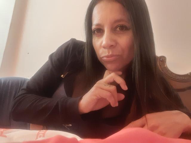 IsabellaSweett with long black hair, pouty lips, and serious expression, lying on bed, wearing black top, hand under chin, red blanket in foreground, brown headboard in background.