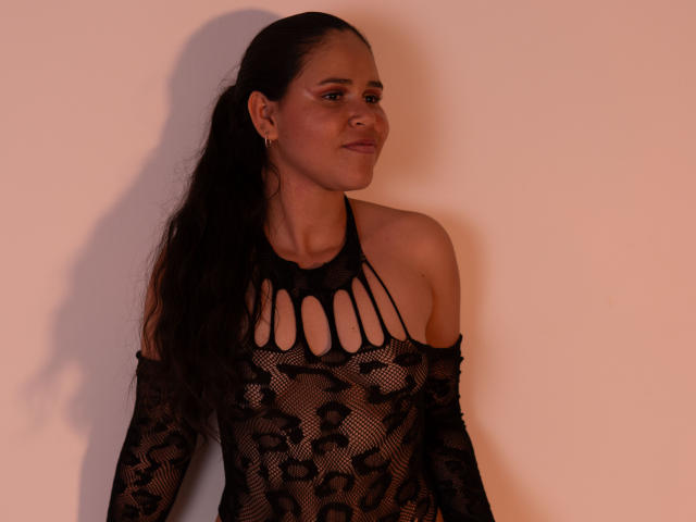 DanTaylor with long black hair, wearing a black lace top with cut-out neck, exposing small tits, standing against a pinkish wall, looking to the side, smiling slightly.