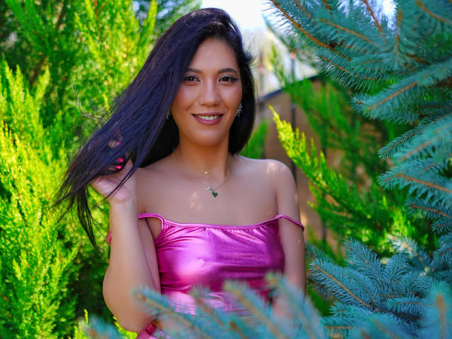 MilaColins with long black hair, wearing a shiny pink off-shoulder top, smiling, standing amidst green pine trees, touching her hair with one hand. Her tits are small.