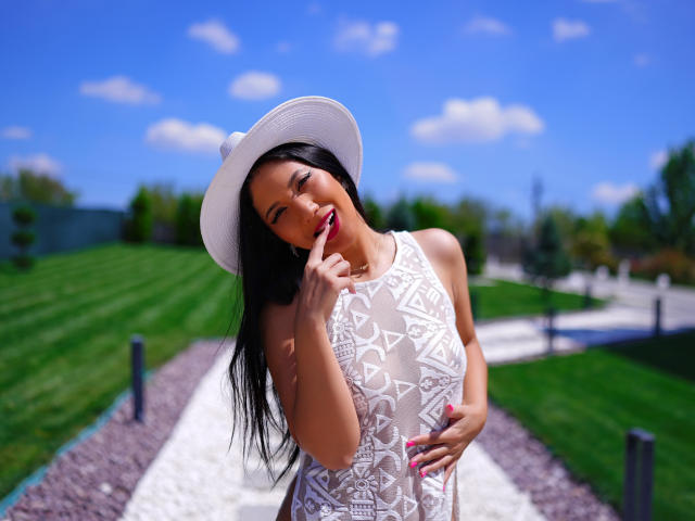 MilaColins with long black hair, wearing a white hat and lace dress, touching her pink-lipsticked lips, hand on her chest, blue sky and green grass in the background.