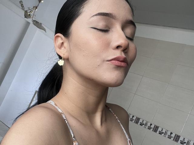 DeisyWest with closed eyes, black eyeliner, pink lips, small zit on cheek, wearing floral bra, dark ponytail, white heart earrings, light skin, subtle makeup.