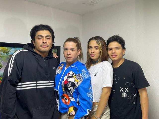 Four TsWomnTwoMenAnJoy stand in a room, wearing casual clothes. One has black hair and a hoodie, another in a blue shirt with a comic print, and two in black shirts.