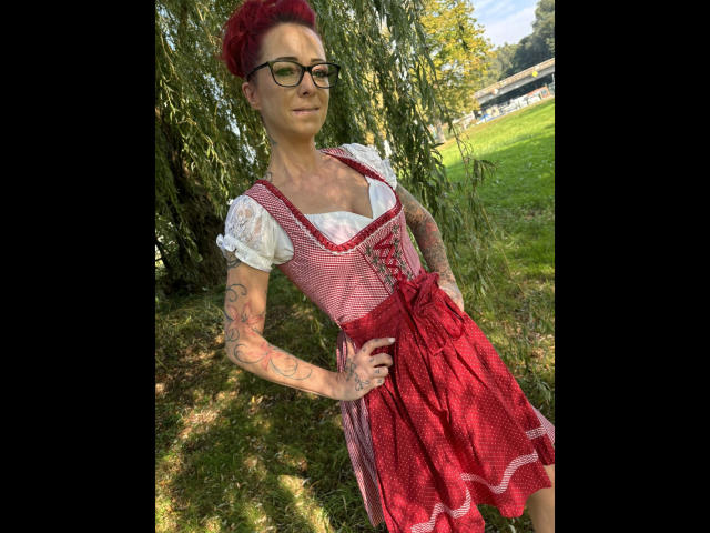 SissySexy-hot with red curly hair, glasses, and tattoos, wearing a red and white German-style dress, standing in a grassy area, hands on hips, smiling. Boobs visible.