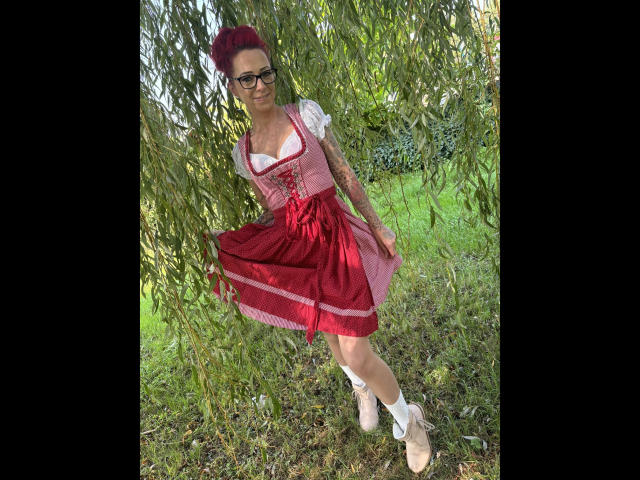 SissySexy-hot with red hair, glasses, tattoos, wearing a pink dress with red ribbon, white puffed sleeves, and pink ballet flats, standing under green willow branches.