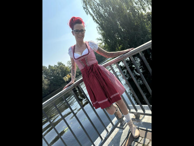 SissySexy-hot with red hair, tattoos, glasses, white shirt, pink dress, standing on bridge, holding railing, red shoes, arms out, sunny, water, trees in background.