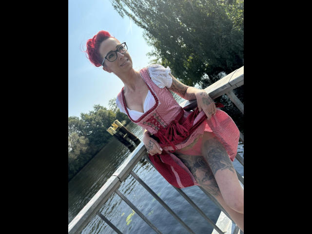 SissySexy-hot with red hair, glasses, and tattoos, wearing a pink dress with white blouse, standing on a bridge, exposing her tits and tattooed thighs, against a sunny backdrop.