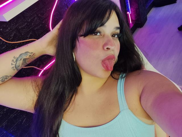 AntonelaPark has long black hair, puffy pink lips, and a tattoo on her right arm. She's sticking her tongue out, wearing a blue tank top, and gold hoop earrings.