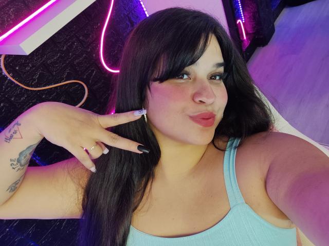 AntonelaPark with long black hair, pink lipstick, light blue tank top, showing off tattoos on her hand and upper arm, making a peace sign, under purple neon lights.