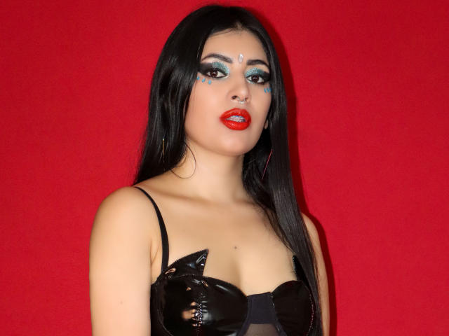 AllisonMuller with long black hair, red lipstick, blue eye makeup, black strapless top with shiny black cups, red background, looking at camera. Boobs visible, nose pierced.