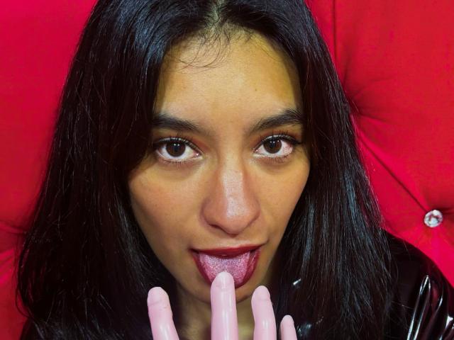 VennusGhirls with dark hair, brown eyes, and tan skin, licking a pink dildo with her tongue out, wearing a black latex top. Red background.