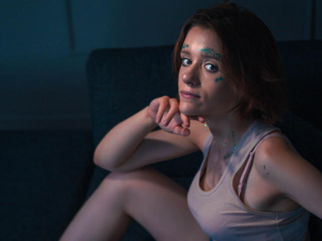 FrancesaL with short brown hair, teal face paint, and a pink tank top, sitting on a dark blue couch, smiling with her chin on her hand, looking at the camera.